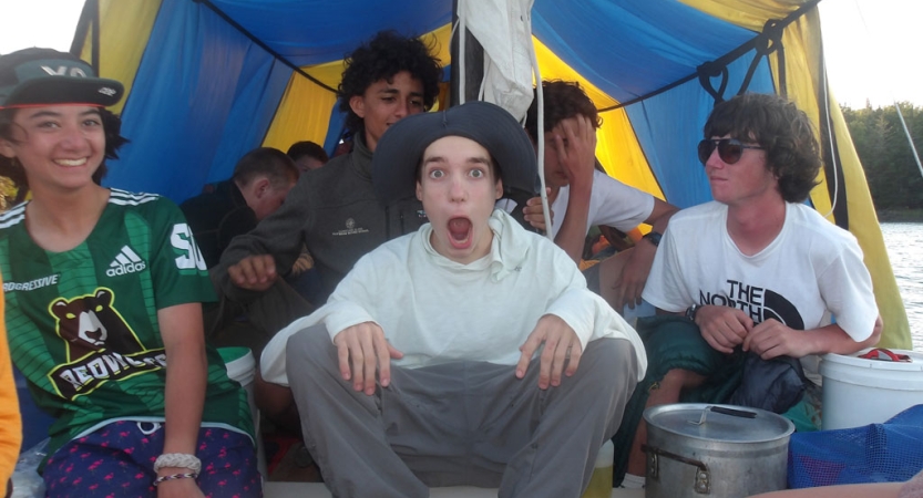 A group of students sit under a tarp on a sailboat. One of them makes a funny face. 